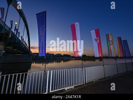 01 maggio 2024, Brandeburgo, Francoforte (Oder): Le bandiere ondeggiano al vento all'alba di fronte al ponte cittadino sul fiume Oder tra Francoforte (Oder) e Slubice in Polonia. In occasione del ventesimo anniversario dell'adesione della Polonia all'Unione europea, i ministri degli Esteri dei due paesi vicini vogliono riunirsi a mezzogiorno. Entrambi vogliono visitare il Collegium Polonicum a Slubice sul lato polacco, nonché un festival europeo, e poi camminare insieme attraverso il ponte della città sul confine tedesco-polacco fiume Oder fino a Francoforte (Oder). Foto: Patrick Pleul/dpa Foto Stock