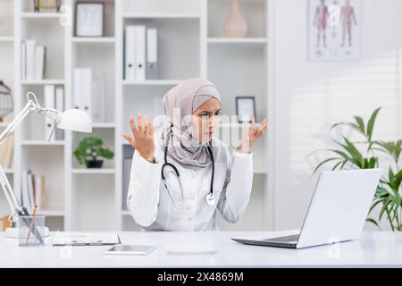 Una donna professionista che indossa un hijab sembra frustrata mentre lavora al suo laptop in un moderno ufficio clinico. Immagine perfetta per le sfide del settore sanitario. Foto Stock