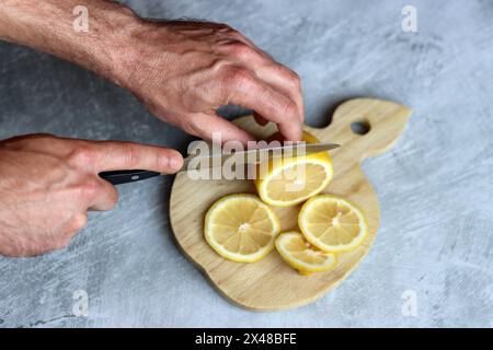 Mani maschili che tagliano il limone su una tavola di legno. Sfondo grigio con spazio di copia. Foto di preparazione della limonata. Foto Stock