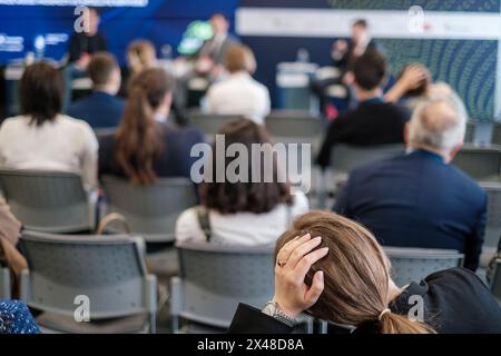 Immagine mirata di una donna che si sente stressata o sopraffatta mentre partecipa a un seminario aziendale. Si siede tra i membri del pubblico sfocati in un ambiente moderno per conferenze. Foto Stock