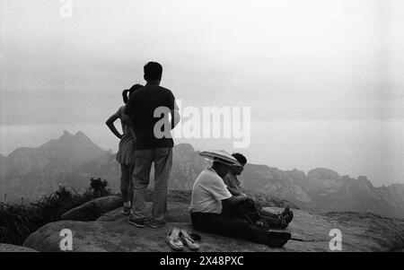 Nell'estate del 2014, in cima al monte Lao a Qingdao, nella provincia di Shandong, Un piccolo gruppo di individui si erge e siede in cima a una montagna rocciosa, che si affaccia su una vasta distesa di montagne scoscese sotto un cielo nebbioso. L'atmosfera è contemplativa e serena, mentre ammirano la bellezza naturale del paesaggio a fine giornata. /Cina Foto Stock