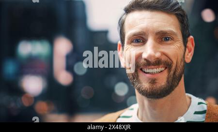 Ritratto di un uomo barbuto felice e bello in abiti casual in piedi in strada guardando la fotocamera, sorridente. Elegante giovane adulto o maschio di mezza età con barba. Sfocata città di scavo sullo sfondo Foto Stock
