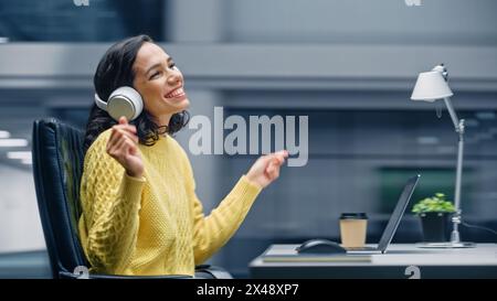 Ufficio moderno: Donna d'affari ispanica sorridente seduta alla scrivania che lavora al computer, indossa le cuffie, ascolta musica, balli. E' felice, si diverte, divertirsi. Sfondo sfocatura movimento Foto Stock