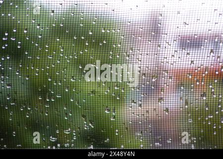 L'acqua piovana cade sulla finestra di garza con lo sfondo di piante e cappotti Foto Stock