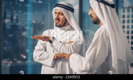 Due uomini d'affari musulmani di successo in White Traditional outfit in piedi in ufficio riuniscono, negoziano e parlano di opportunità finanziarie. Arabia Saudita, Emirati, concetto di uomo d'affari arabo. Foto Stock