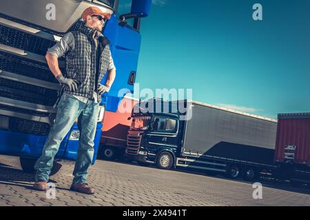 Autista di semi-camion europeo caucasico che soggiorna accanto al suo semi-camion blu. Tema lavoro camionisti. Foto Stock