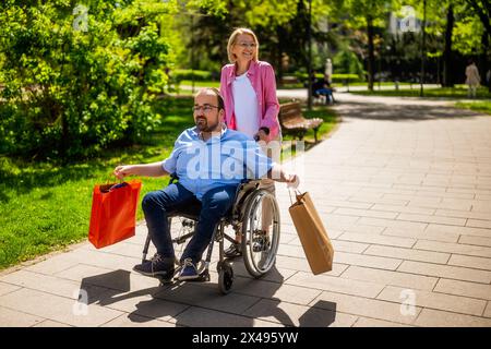 L'uomo in sedia a rotelle sta andando dallo shopping. Il suo amico lo sta aiutando con la sedia a rotelle. Foto Stock