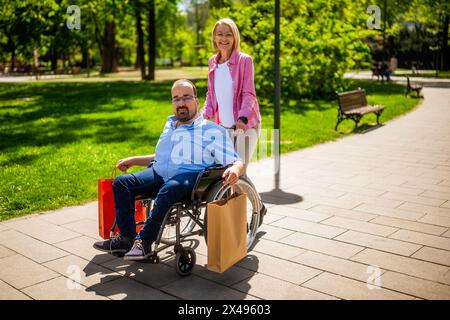 L'uomo in sedia a rotelle sta andando dallo shopping. Il suo amico lo sta aiutando con la sedia a rotelle. Foto Stock