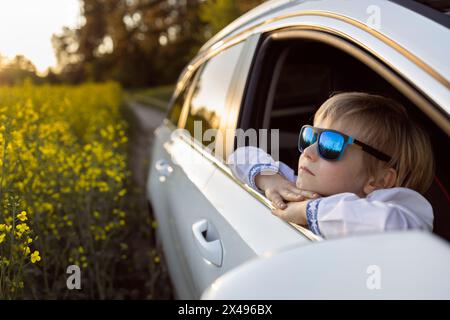 la faccia felice di un bambino con gli occhiali da sole blu guarda fuori dal finestrino dell'auto aperto e si guarda intorno con interesse. viaggi in auto con la famiglia in estate Foto Stock