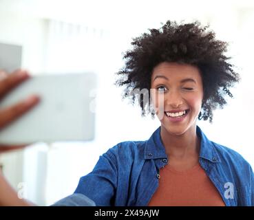 Ritratto, donna e selfie con l'occhiolino in casa per social media, immagine del profilo ed espressione divertente. Donna, smartphone e emoji lampeggiante per Foto Stock