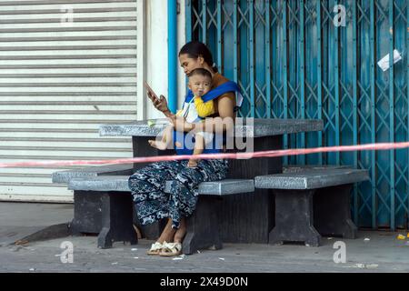 SAMUT PRAKAN, THAILANDIA, 20 aprile 2024, Una donna siede con un bambino tra le braccia vicino ai negozi chiusi sulla strada Foto Stock