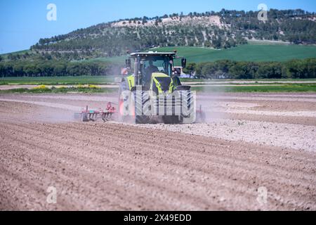 Un agricoltore con il suo trattore che trasporta un aratro che rastrella grandi campi di raccolto e solleva molta polvere mentre lo fa con una collina piena di alberi nella schiena Foto Stock