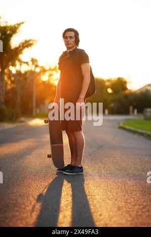 Ritratto, uomo e skateboard in esterni, strada e zaino con cuffie, estate e tramonto. Studente universitario, attività fisica e sport in Foto Stock
