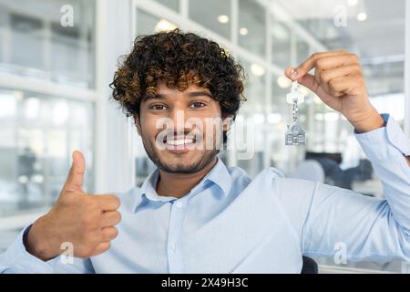 Diversi agenti immobiliari sorridono alla telecamera mentre mostrano un gesto con il pollice in alto e tengono le chiavi con una catena a forma di casa. Un uomo cordiale consiglia di acquistare un appartamento in un nuovo complesso residenziale. Foto Stock