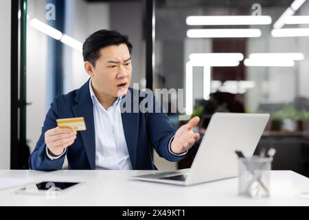 Uomo asiatico confuso in camicia e giacca che guarda lo schermo del computer e tiene in mano la carta di credito. Uomo adulto arrabbiato che cerca di aumentare il limite di credito sul sito web della banca e riceve risposta di rifiuto. Foto Stock