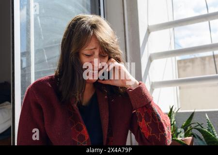 Donna latina adulta seduta sul balcone di casa sua piangendo triste parlando al telefono con il suo ragazzo. Foto Stock