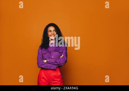 Donna felice con i capelli ricci, in abbigliamento casual, si erge con cura in un vivace studio. Il suo sorriso brillante e la sua espressione pensiva rivelano la sua elegante a Foto Stock