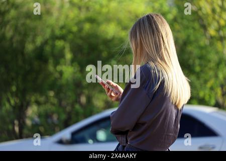 Ragazza con lunghi capelli biondi che usa lo smartphone per le strade cittadine sullo sfondo sfocato dell'auto Foto Stock