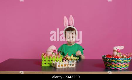 Bambino piccolo felice che organizza uova e pulcino in un cestino in preparazione alle celebrazioni della domenica di pasqua, creando decorazioni natalizie in studio. Bambino che si diverte a colorare. Telecamera B.. Foto Stock