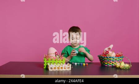 Piccolo ragazzo allegro che crea decorazioni pasquali fatte a mano dipingendo con acquerelli e pennelli. Bambino piccolo che usa forniture d'arte colorando uova per le vacanze e decorando attività. Telecamera B.. Foto Stock