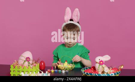 Bambino piccolo felice che organizza uova e pulcino in un cestino in preparazione alle celebrazioni della domenica di pasqua, creando decorazioni natalizie in studio. Bambino che si diverte a colorare. Telecamera A.. Foto Stock