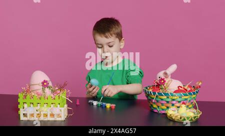 Gioioso bambino che dipinge uova per le festività natalizie di pasqua in studio, utilizzando acquerelli e forniture d'arte. Sorridente colorazione in età prescolare ornamenti festivi in preparazione per la domenica. Telecamera A.. Foto Stock