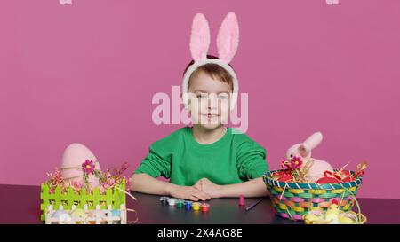 Bambino piccolo che mette le orecchie da coniglio sulla testa in studio, preparandosi per le festività della domenica di pasqua. Allegro bambino seduto a un tavolo per dipingere le uova e creare ornamenti natalizi. Telecamera A.. Foto Stock