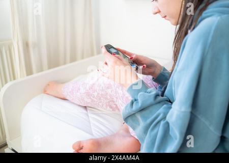 Una giovane donna guarda il suo telefono cellulare che scorre sullo schermo con unghie dipinte di rosso. È seduta a piedi nudi su un letto, con luce soffusa che arriva in ge Foto Stock