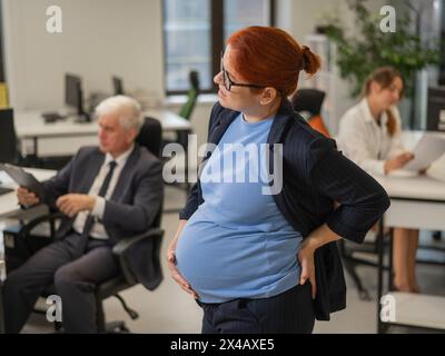 Una donna incinta soffre di dolore e tiene lo stomaco in piedi nel mezzo dell'ufficio accanto ai suoi colleghi. Foto Stock