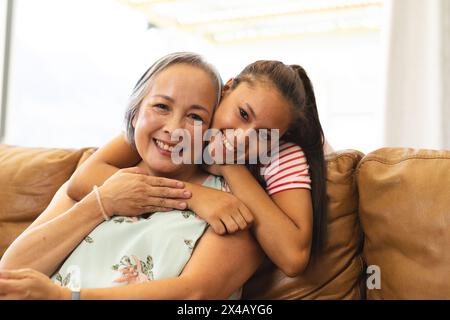 Nonna asiatica e nipote adolescente birazziale che si abbraccia sul divano a casa. Indossando camicie floreali e a righe, condividendo sorrisi e godendosi il calore Foto Stock