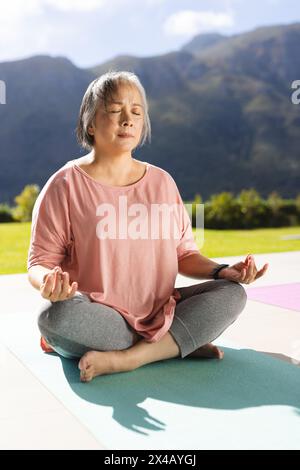 Donna asiatica anziana seduta a gambe incrociate su tappetino per yoga, meditando all'aperto. Ha i capelli grigi corti, gli occhi chiusi e indossa abiti da allenamento casual, unal Foto Stock