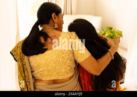 Madre indiana e figlia adolescente che si abbracciano calorosamente a casa. Entrambi indossano abiti indiani tradizionali, mostrando amore e vicinanza, inalterati Foto Stock