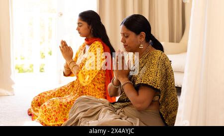 Madre indiana e figlia adolescente, seduta a zampe a casa, pregando insieme. Entrambi con lunghi capelli neri, con la madre che mostra segni di invecchiamento, Foto Stock