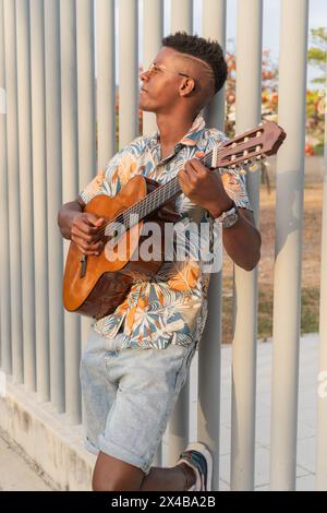 Un giovane africano si appoggia contro una recinzione metallica, perduta nella musica mentre suona la sua chitarra, indossando una camicia tropicale e occhiali da sole durante il tramonto. Foto Stock