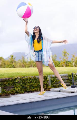 Giovane donna birazziale che gioca con il Beach ball all'aperto a bordo piscina. Indossando un top giallo, pantaloncini in denim e lunghi capelli neri, si gode il sole Foto Stock