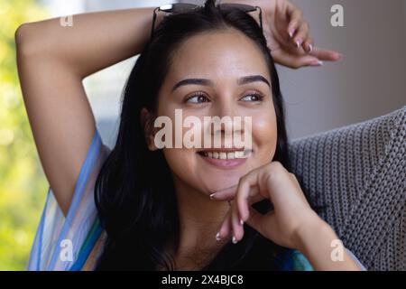 Giovane donna birazziale che si rilassa sul divano a casa, guardando lontano. Ha lunghi capelli scuri, pelle marrone chiaro, e indossa uno scialle colorato, inalterato Foto Stock