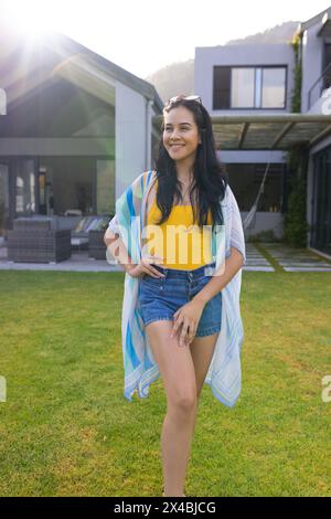Giovane donna birazziale in piedi all'aperto in un cortile soleggiato. Indossando un top giallo e pantaloncini in denim, tenendo lo scialle leggero, godendosi la calda luce del sole, inalterato Foto Stock