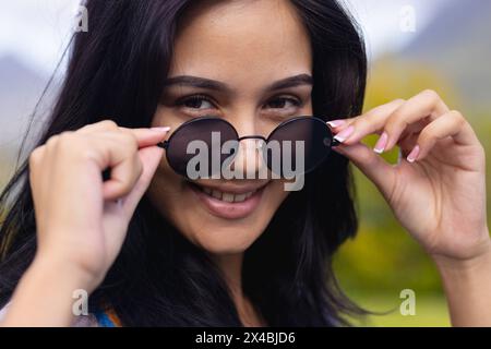 Giovane donna birazziale con occhiali da sole, sorridente all'aperto a casa. Ha lunghi capelli scuri, trucco chiaro e smalto rosa, inalterato Foto Stock