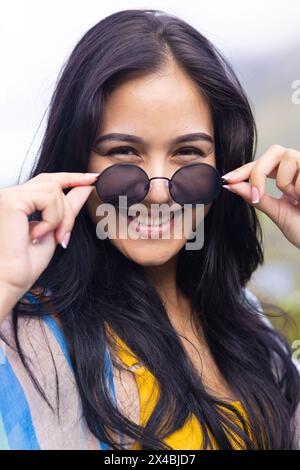 Giovane donna birazziale che tiene gli occhiali da sole e sorride alla macchina fotografica all'aperto. Indossando un top colorato, presenta lunghi capelli scuri, godendosi la giornata di sole, unaltere Foto Stock