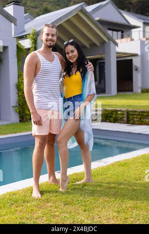 Coppia variegata in piedi all'aperto vicino alla piscina, sorridendo davanti alla macchina fotografica. Lui, con pelle leggera e capelli biondi corti; lei, con pelle d'oliva e lunghi capelli neri, enj Foto Stock
