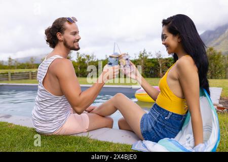 Donna birazziale e uomo caucasico che si godono un drink all'aperto a bordo piscina. Entrambi hanno i capelli scuri, lei con i capelli lunghi e dritti, lui indossa gli occhiali da sole, ridi Foto Stock