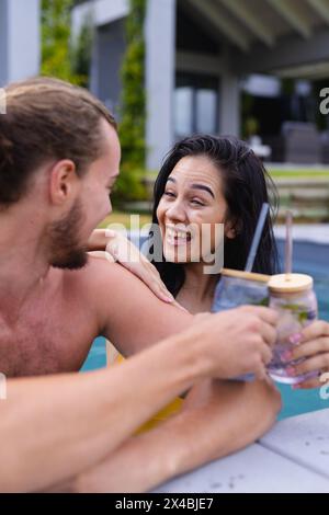 Coppie diverse che si divertono all'aperto in piscina, ridendo insieme. Donna birazziale con lunghi capelli neri che tiene in mano una bevanda, di fronte all'uomo caucasico, entrambi più affilati Foto Stock