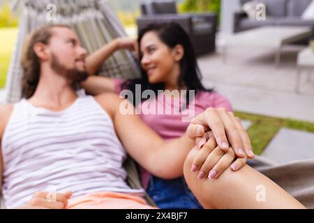 Coppie diverse che si rilassano all'aperto vicino alla piscina, tenendosi per mano. Donna birazziale con capelli scuri e uomo caucasico con barba e capelli lunghi, che condivide la tranquilla momen Foto Stock