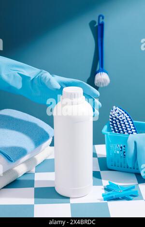 Una mano con guanti di gomma si prepara a raccogliere un flacone bianco di detergente senza etichetta. Prodotti per la pulizia posizionati su un quadratino blu e bianco Foto Stock