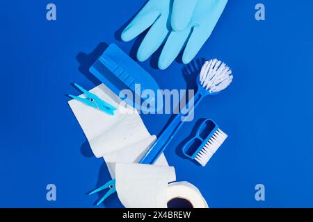 Set di materiali di consumo per la pulizia su sfondo a colori. Vista dall'alto di spazzole, guanto in gomma e rotolo di carta decorati su sfondo blu. Servizio di pulizia della casa e Foto Stock