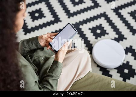 Vista ad angolo alto di una giovane donna irriconoscibile che utilizza l'app per aspirapolvere robotizzato su smartphone Foto Stock