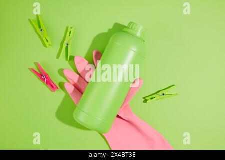 Prodotti per la pulizia colorati su sfondo verde semplice. Un guanto in gomma rosa contenente una bottiglia verde di detergente senza etichetta per il design. Vista dall'alto, Hous Foto Stock