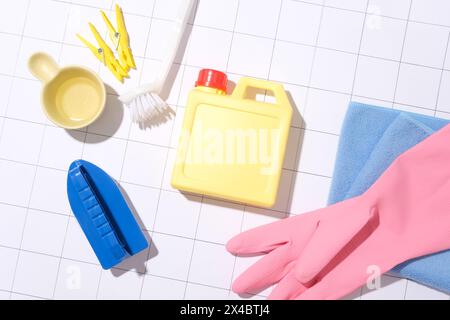 Una bottiglia di detergente gialla senza marchio, circondata da una spazzola, guanti di gomma, asciugamano di cotone, clothespin e paletta per l'acqua su un backgroun bianco a scacchi Foto Stock