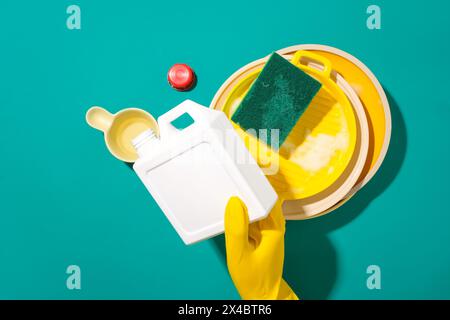 Una mano che indossa un guanto di gomma giallo tiene una bottiglia di detergente e la versa in una paletta per l'acqua, accanto a una spugna con piatti sporchi. Copia spazio per A. Foto Stock