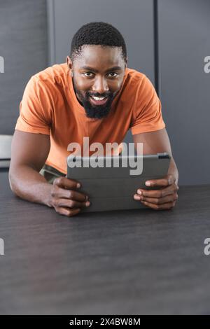 A casa, proprietario di casa afroamericano maschio sdraiato, tenendo un tablet. Indossa una camicia arancione che mostra un sorriso amichevole e rilassati con un backgroun blu Foto Stock